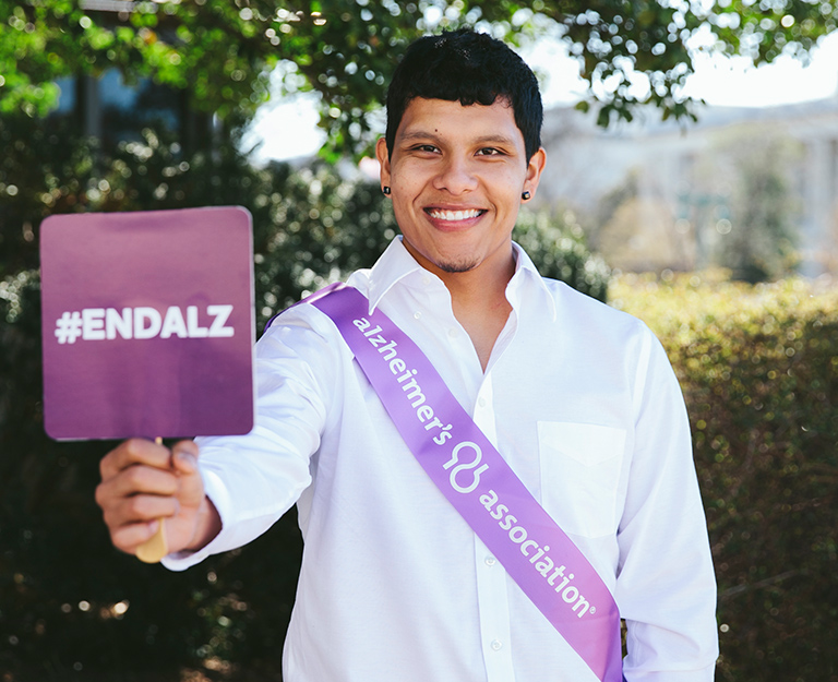 Image for Alzheimer's and Brain Awareness Month - Young man standing in front of trees and bushes holding a purple sign, #ENDALZ. He is wearing a white button up and a Alzheimer's Association sash.