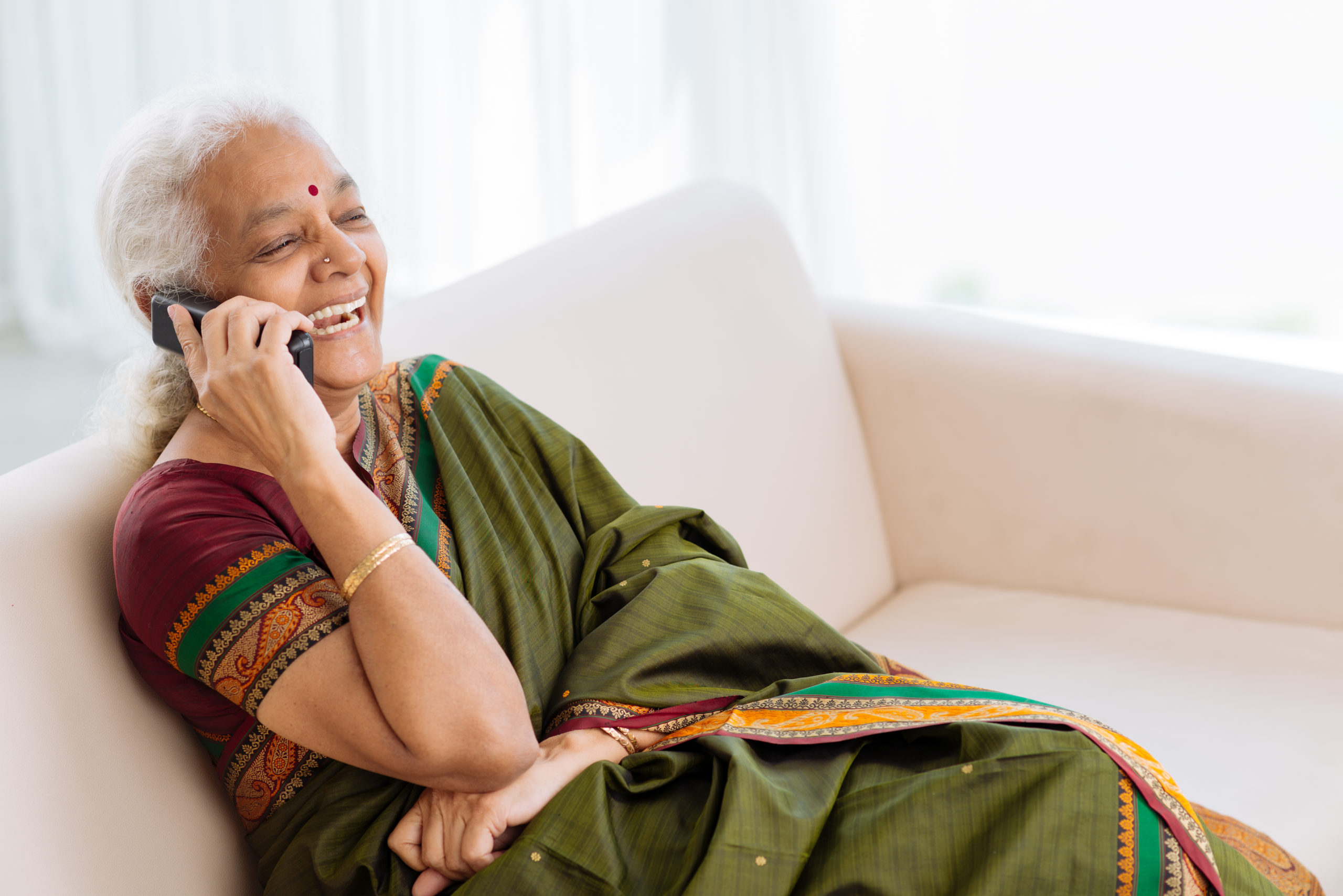 Laughing Indian woman with a red bindi wearing a beautiful green and red decorated sari sits on a cream colored sofa and talks on the telephone. Behind her there are white shades and light shining through.