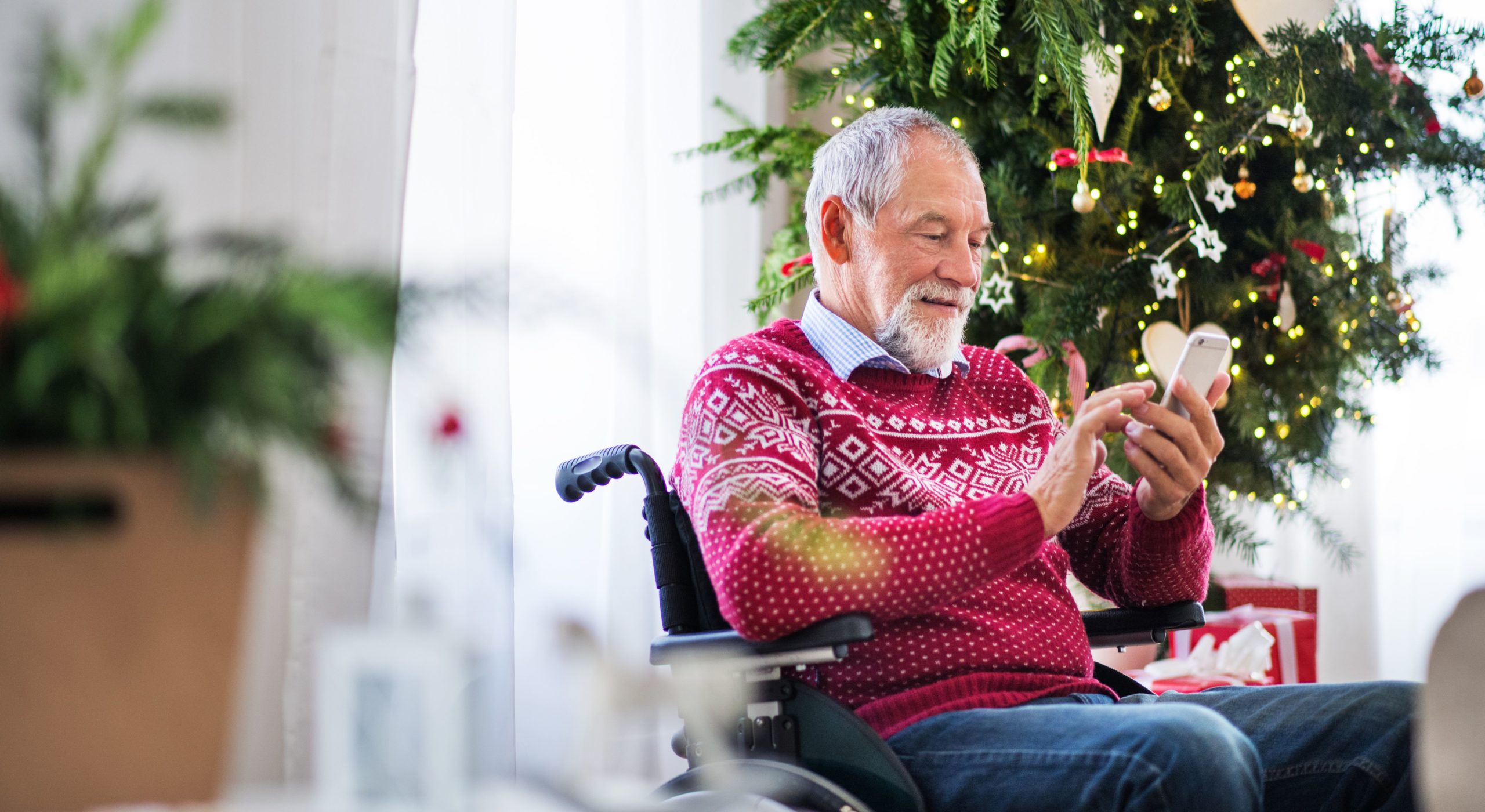 A senior man wearing a red sweater sits in a wheelchair and texts on his phone at home during the holidays. Behind him is a Christmas tree with gifts below, and white curtains with light shining through.